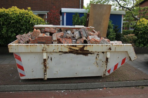 A large metal skip container positioned on a street curb in Sutton, filled with a mixture of red brick debris and small concrete pieces, some of which are uneven and broken, indicating recent demolition or clearance work. The skip shows signs of rust and weathering, with visible streaks of rust across its white painted surface, and red and white striped reflective tape along its sides for safety. Behind the skip, there is a background of residential buildings, including a blue shed or garage, and surrounding greenery such as bushes and small trees, suggesting the scene is within a suburban garden clearance operation managed by Garden Clearance Sutton. The street is paved with red and grey bricks, and the overall setting appears to be during daylight hours with natural lighting highlighting the textures and colors of the debris and surroundings.