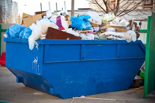 A large, blue industrial rubbish skip filled with various types of waste, including cardboard boxes, plastic bags, white foam packaging, and miscellaneous debris, situated on a paved surface during daylight. The skip is overflowing with recyclable and non-recyclable rubbish, with some material piled higher than the rim. In the background, there are glimpses of trees and a building, indicating an outdoor urban or suburban environment, possibly in Sutton. The scene appears to be part of a professional waste removal or garden clearance service, as referenced by the website gardenclearancesutton.co.uk, highlighting services related to rubbish collection and clearance. The overall environment is clean with no immediate signs of disposal activity, emphasizing the static state of the waste container for collection or disposal. The image reflects the typical appearance of waste removal operations in a residential or commercial setting within the local area.