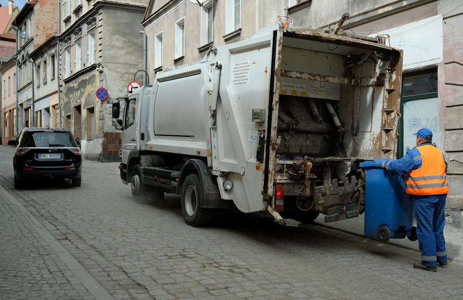 Close-up of a person wearing a yellow and grey checkered shirt, green gloves, and grey trousers, holding open a large black rubbish bag made of thick plastic, which is empty. The person is standing ou