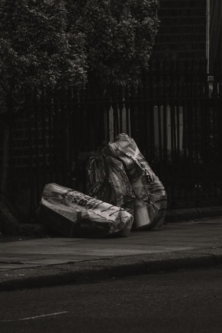 A black wheeled rubbish bin with a white label and the words 'ST. JOHN'S' printed on it is positioned on a paved sidewalk next to a street at night. The bin's lid is open, revealing a variety of disca