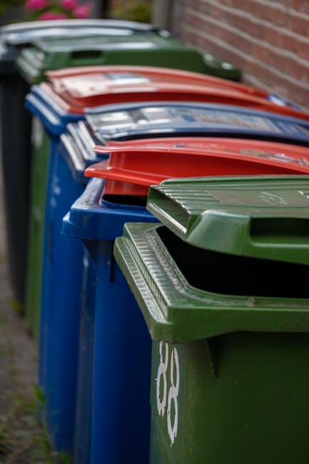 A row of old-fashioned terraced houses with bay windows and slate roofs, situated along a quiet residential street. In the foreground, two black waste bins with slightly weathered surfaces are positio